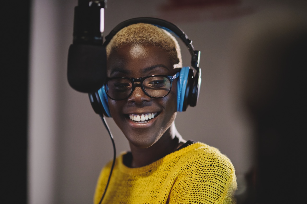 Optimistic ethnic female radio host in bright yellow sweater headphones and glasses recording podcast near microphone in studio